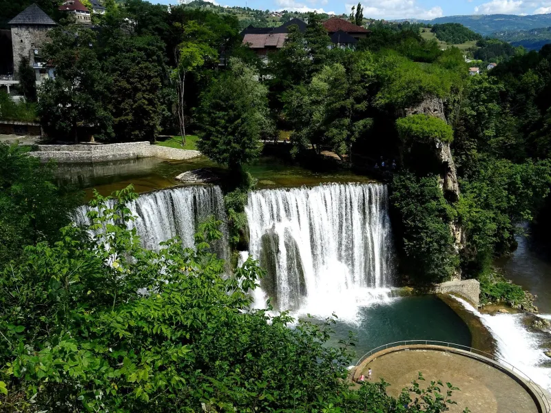The Pliva waterfall in Jajce surrounded by green hills and old town buildings.